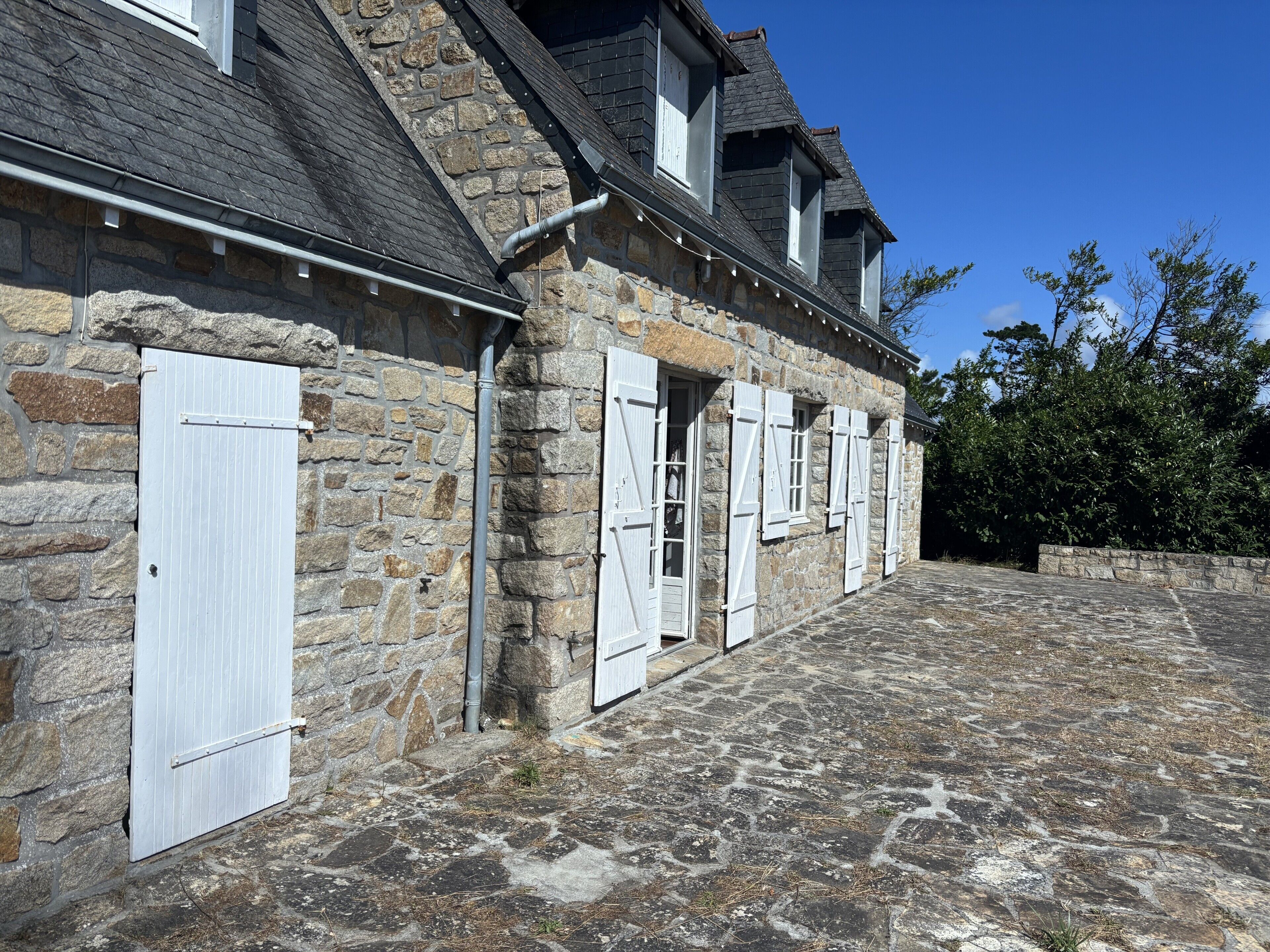 Un edificio de piedra con múltiples ventanas y puertas, un patio empedrado en piedra y un cielo azul despejado.