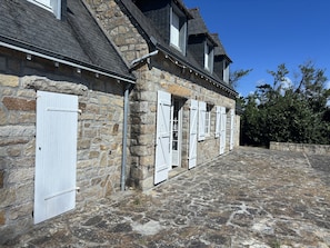 Un edificio de piedra con múltiples ventanas y puertas, un patio empedrado en piedra y un cielo azul despejado.