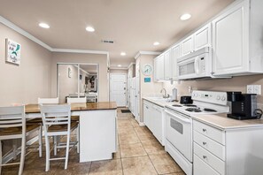 A modern kitchen with white cabinetry, a dining area with a wooden table and chairs, and a visible clock on the wall. - 🐾 Sunset Harbor Palms 2-102 | Pet Friendly Studio on Santa Rosa Sound (Navarre Beach)
