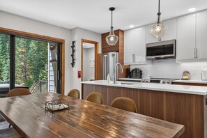 A modern kitchen with white cabinetry, stainless steel appliances, and a wooden dining table. - Walk to Downtown townhome. Private Hot Tub. Views. Sleeps 12 (Winter Park)
