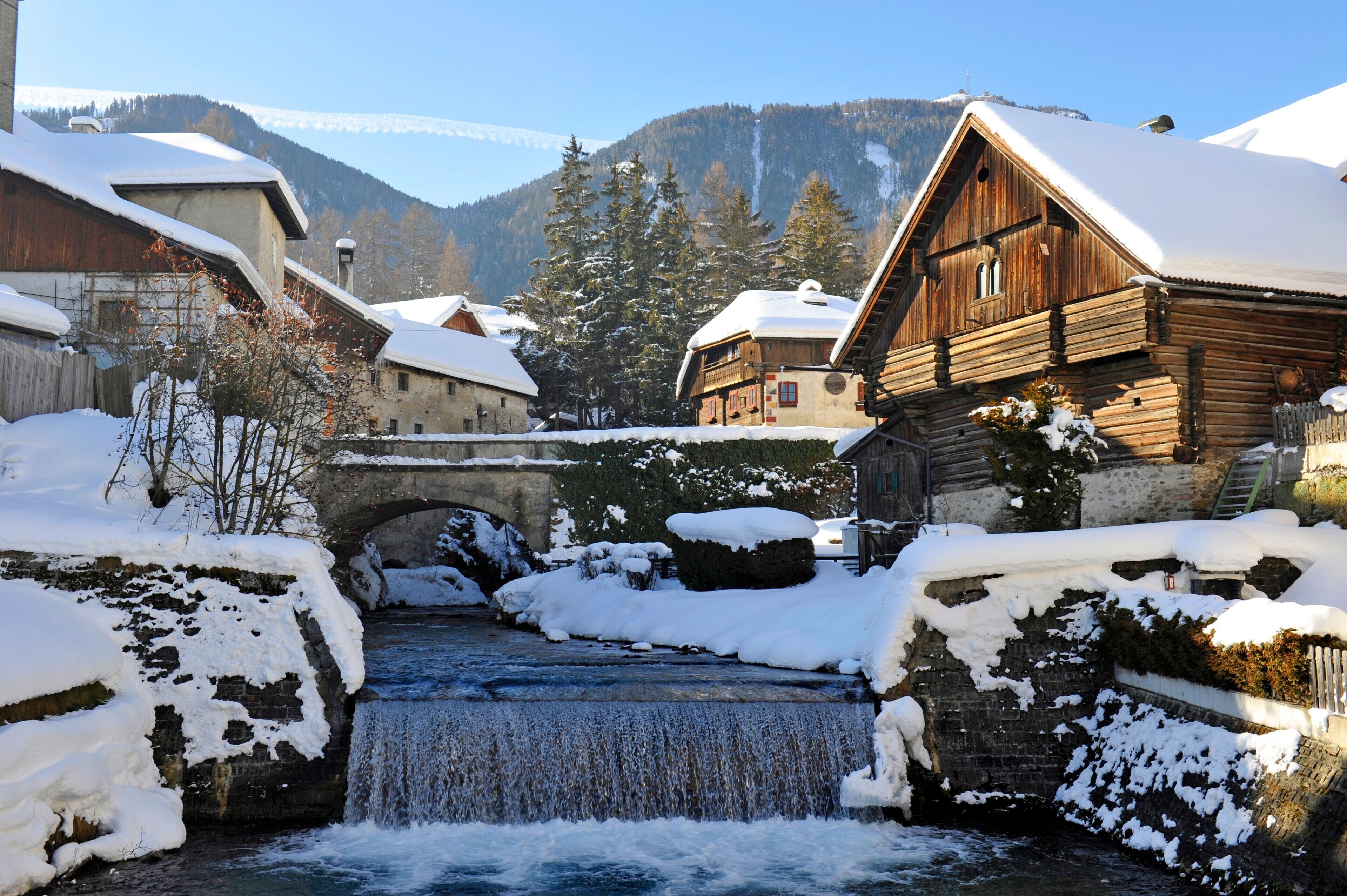 Ein verschneites Dorf mit Holzhäusern, einer Steinbrücke und einem Fluss.