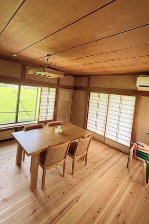 Een kamer met een houten tafel en stoelen, grote ramen met uitzicht op groen, en een plafondventilator.