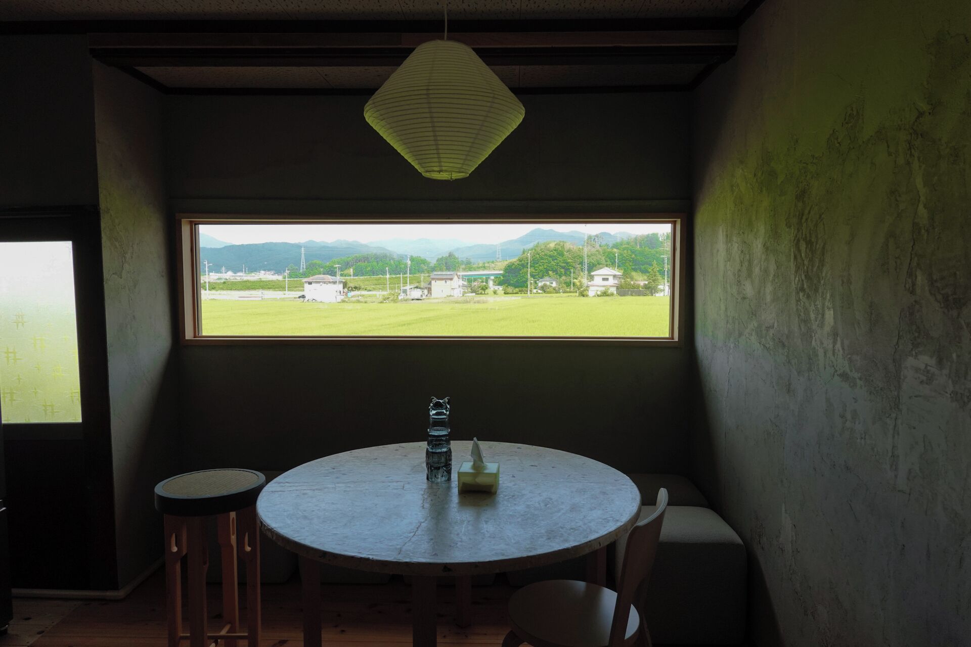 A room with a round table, two chairs, a bottle, and a paper napkin. A window provides a view of a green field and distant buildings.