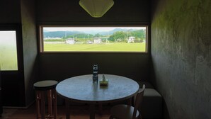 A room with a round table, two chairs, a bottle, and a paper napkin. A window provides a view of a green field and distant buildings.