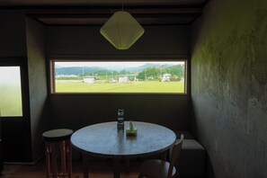 A room with a round table, two chairs, a bottle, and a paper napkin. A window provides a view of a green field and distant buildings.