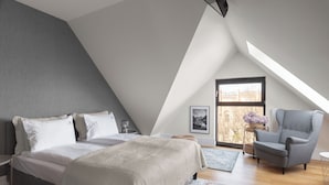 A modern bedroom with a sloped ceiling, a bed with white linens, a grey armchair, and a large window with a view. - Wintera Residence - River View Apartment (Gdańsk)