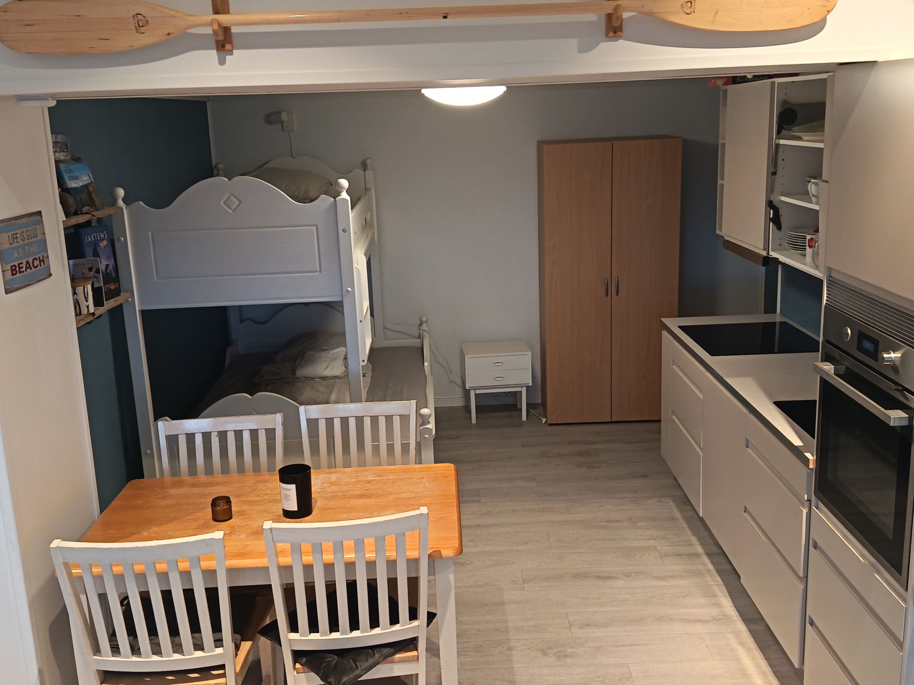 A compact room with a bunk bed, a wooden dining table with chairs, a small bedside table, a wardrobe, and a kitchenette with an oven.
