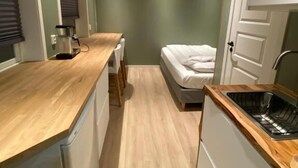 Cozinha compacta com bancada de madeira, pia e uma cama no quarto vizinho.