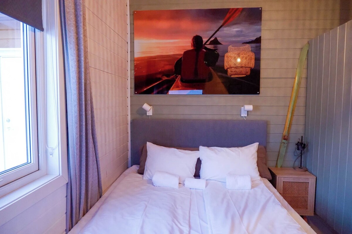 Una habitación de hotel con una cama, un cuadro de un kayakista, una ventana con cortinas y una mesita de noche con una lámpara.
