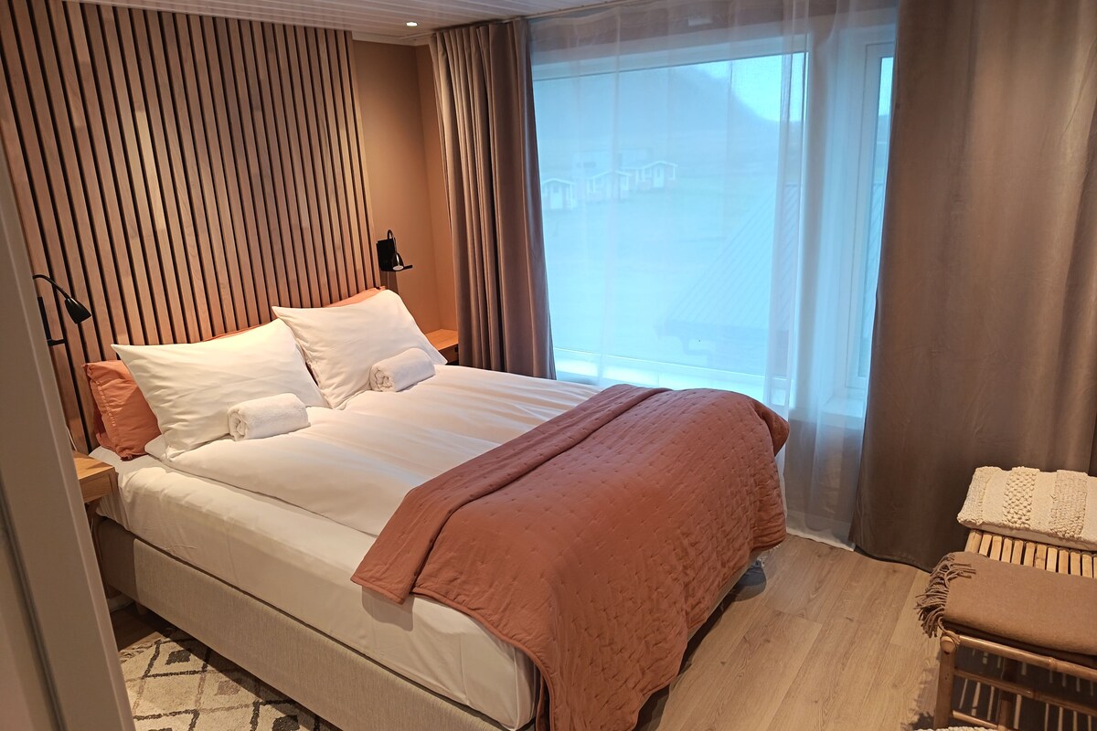 A modern hotel room with a large bed, a bedside table, a lamp, and a view of the outside.