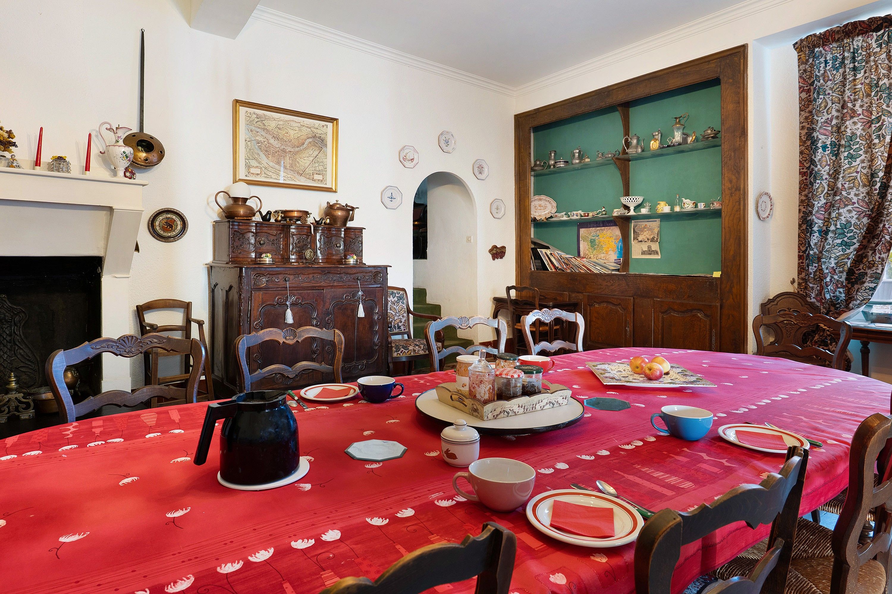 Een eetkamer met een rode tafelkleed, houten stoelen, een open haard en een groene wand met planken.
