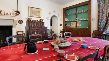 Een eetkamer met een rode tafelkleed, houten stoelen, een open haard en een groene wand met planken.