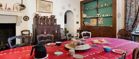 A dining room with a red tablecloth, wooden chairs, a fireplace, and a green wall with shelves.