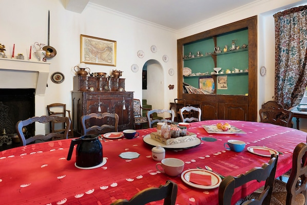 A dining room with a red tablecloth, wooden chairs, a fireplace, and a green wall with shelves.