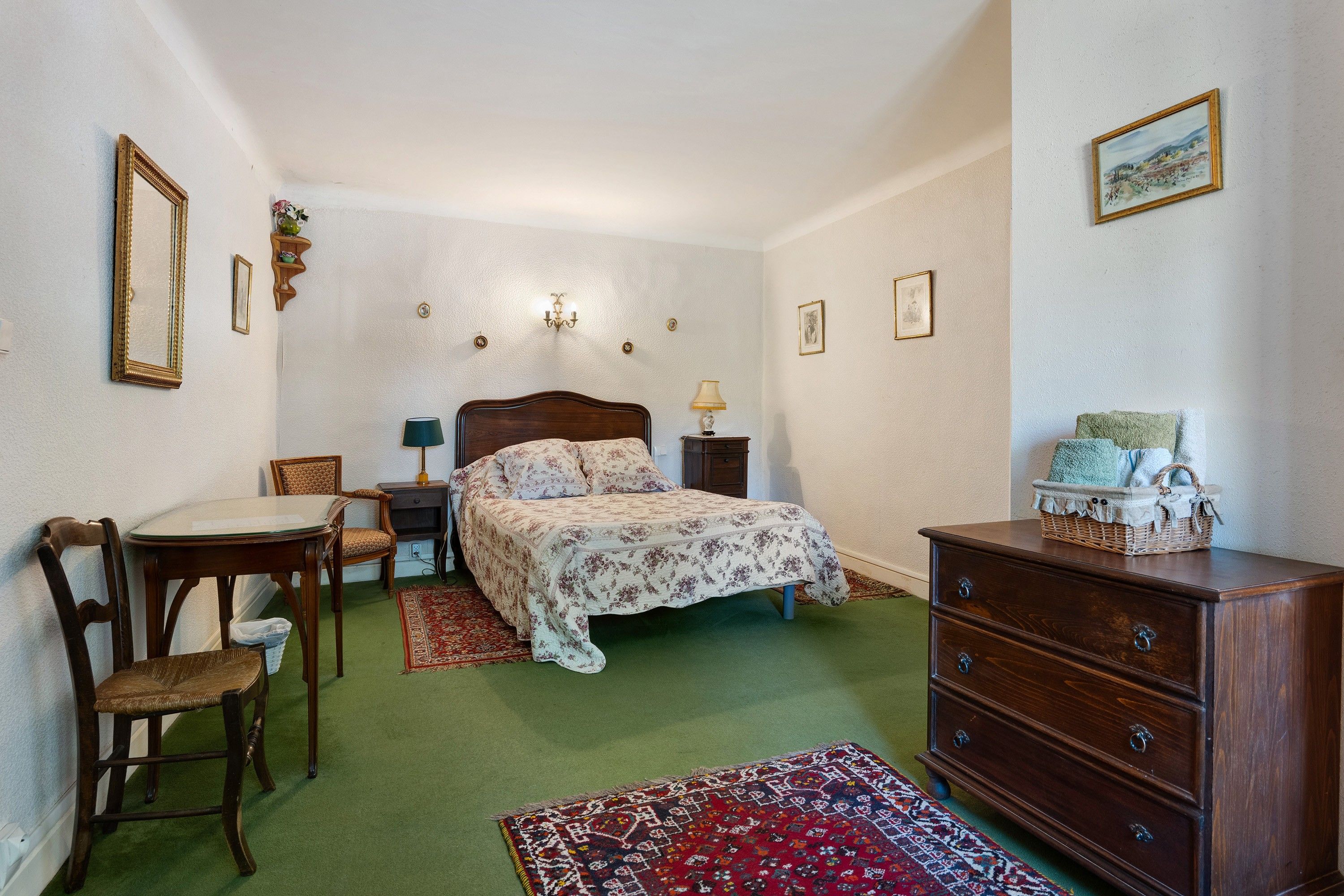 Een slaapkamer met een bed, een dressoir, een stoel, een tafel, een lamp en een vloerkleed.