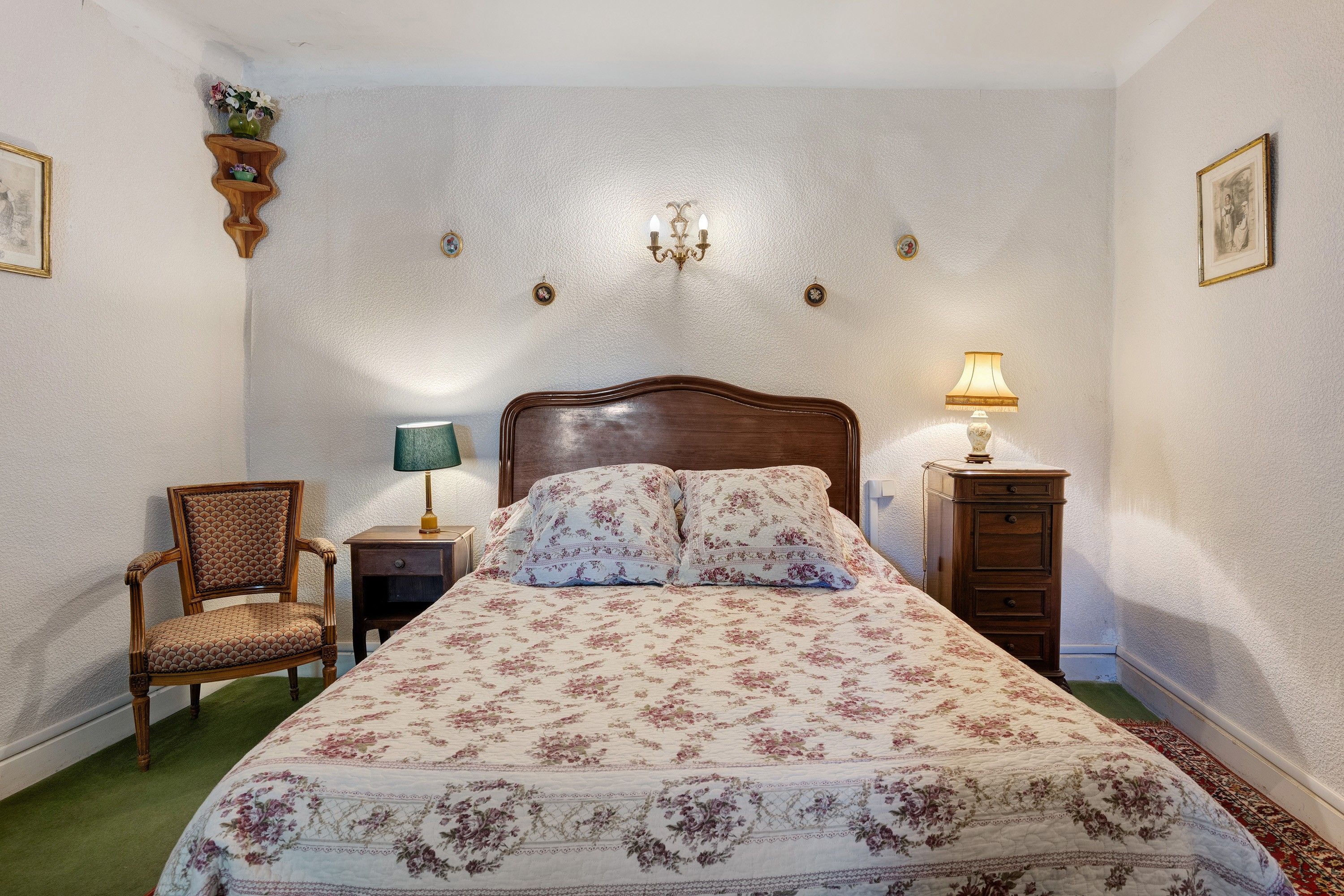 Ein Schlafzimmer mit einem hölzernen Bett, floralem Bettbezug, einem Sessel, einem Nachttisch und einer Wandleuchte.