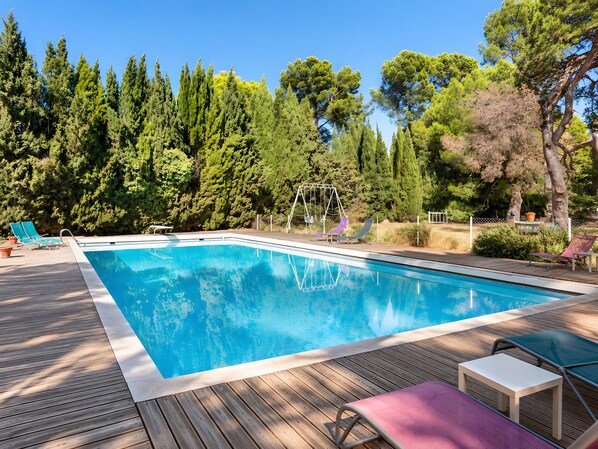 A rectangular swimming pool with a wooden deck, surrounded by greenery and trees.
