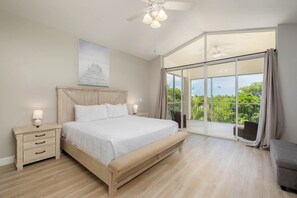 Une chambre dotée d’un grand lit, d’une table de chevet, d’une lampe fixée au mur, d’un ventilateur de plafond et offrant une vue sur la verdure grâce à une porte-fenêtre coulissante.
