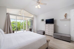 Ein Schlafzimmer mit einem großen Bett, einem Fernseher auf einer Kommode, einem Balkon mit Vorhängen und einem Deckenventilator.