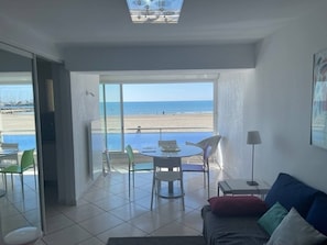 A room with a balcony overlooking a beach, a dining table with chairs, and a sofa with cushions. - Première ligne, t2 cabine pour 4 N123 (Palavas-les-Flots)