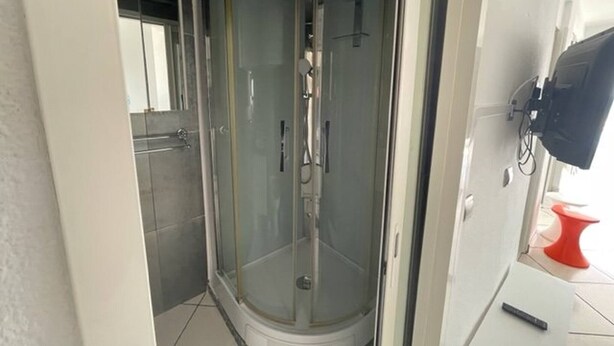 A glass-enclosed shower stall with a tiled floor, a white door, and a red object on the floor. - Première ligne, t2 cabine pour 4 N123 (Palavas-les-Flots)