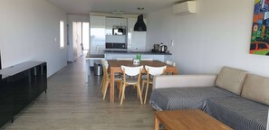A modern living room with a sofa, a dining table with chairs, a kitchen area with a microwave, and a radiator.