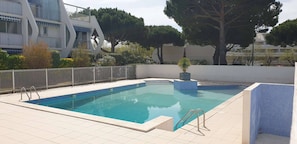 Ein rechteckiger Swimmingpool mit Fliesen und einer umgebenden, gefliesten Terrasse, umgeben von einem weißen Zaun und Bäumen.