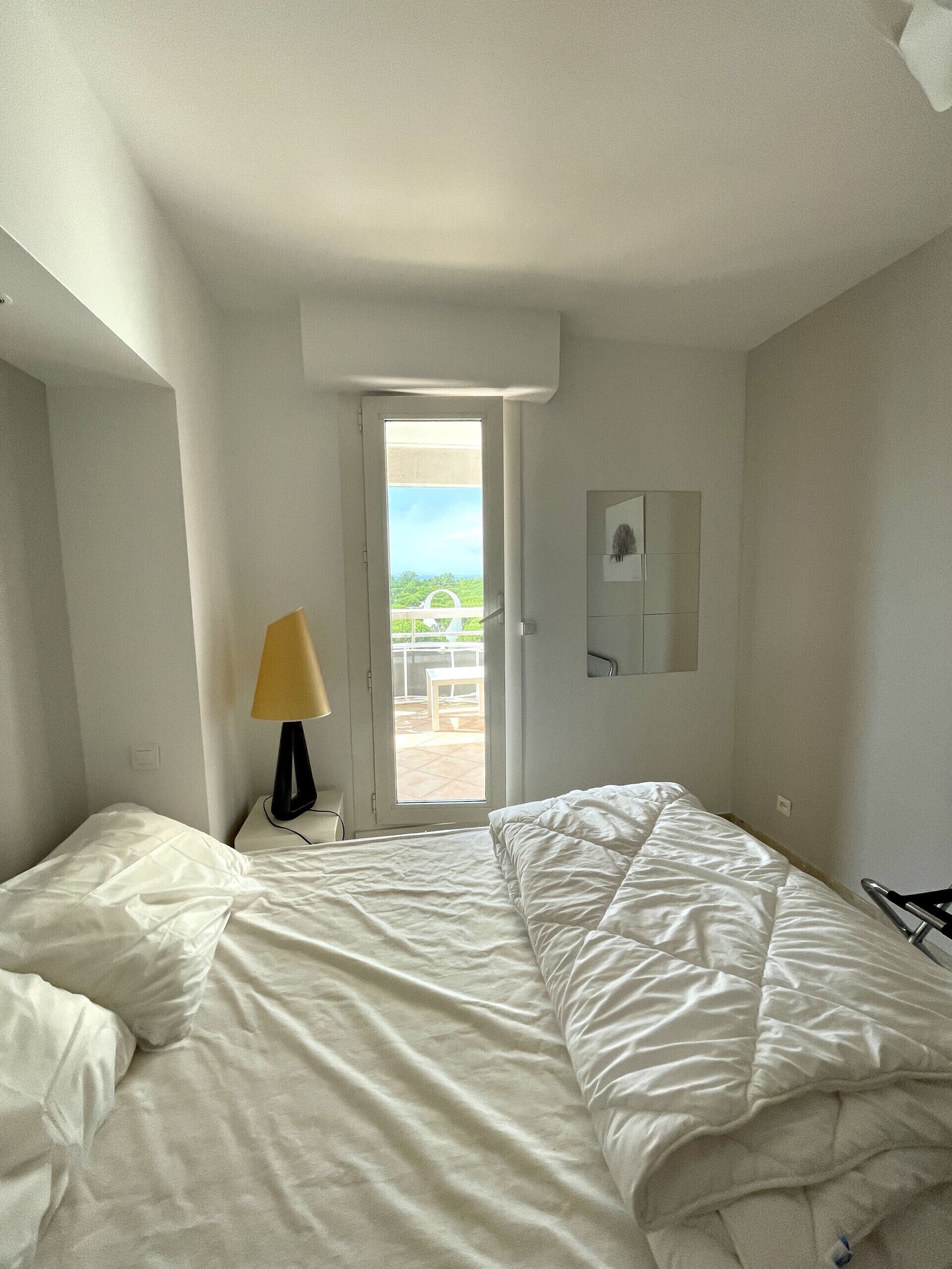 Una habitación de hotel con una cama, una mesita de noche con lámpara, una puerta que da a un balcón y un estante pequeño en la pared.