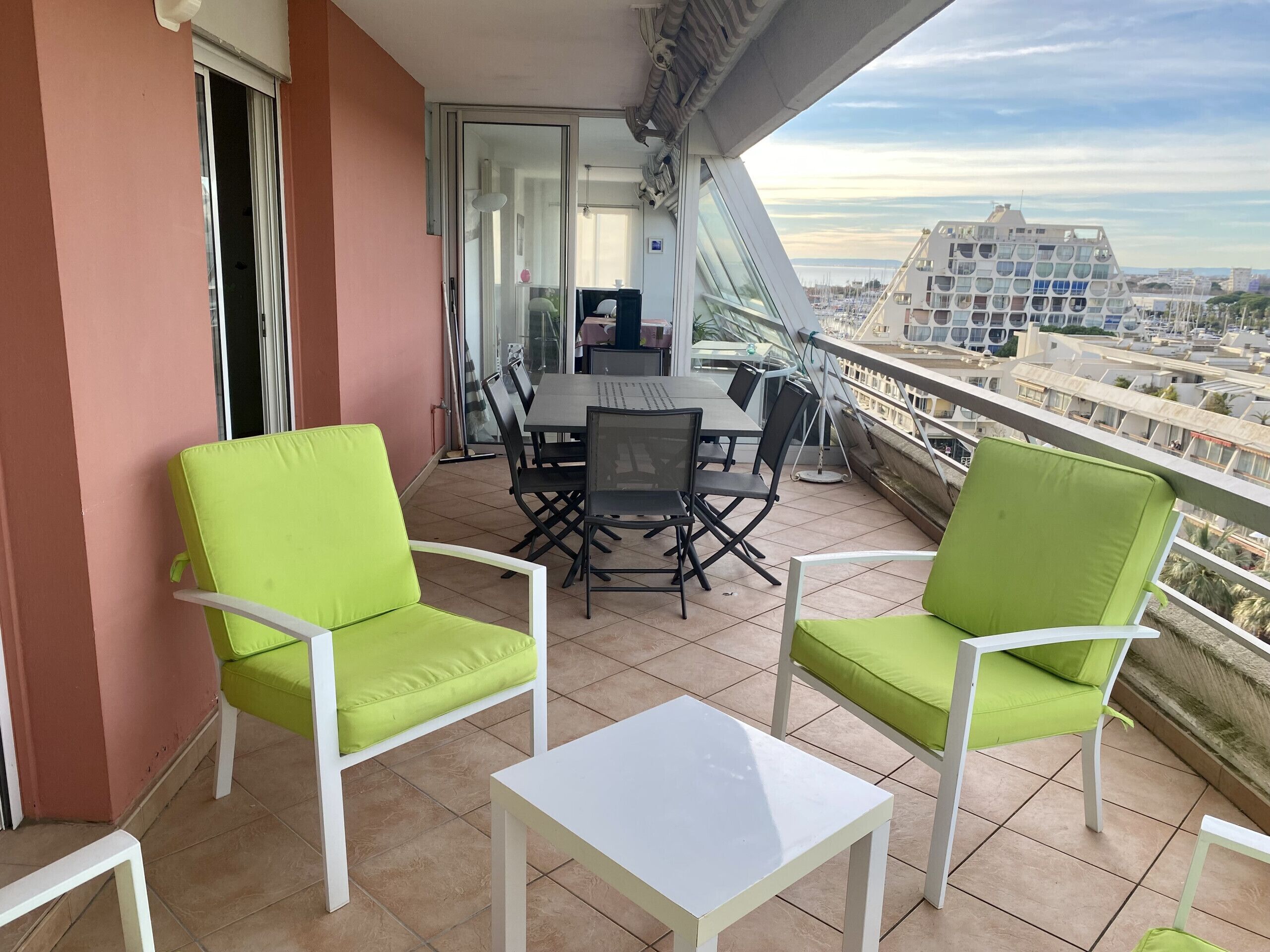 Un balcon avec deux chaises vertes, une table blanche et une porte vitrée donnant sur un espace intérieur.