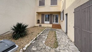 A two-story building with a stone pathway leading to the entrance, a small garden with a plant, and a wooden fence. - Quartier des Goélands, Villa t5 pour 8 N25 (La Grande-Motte)
