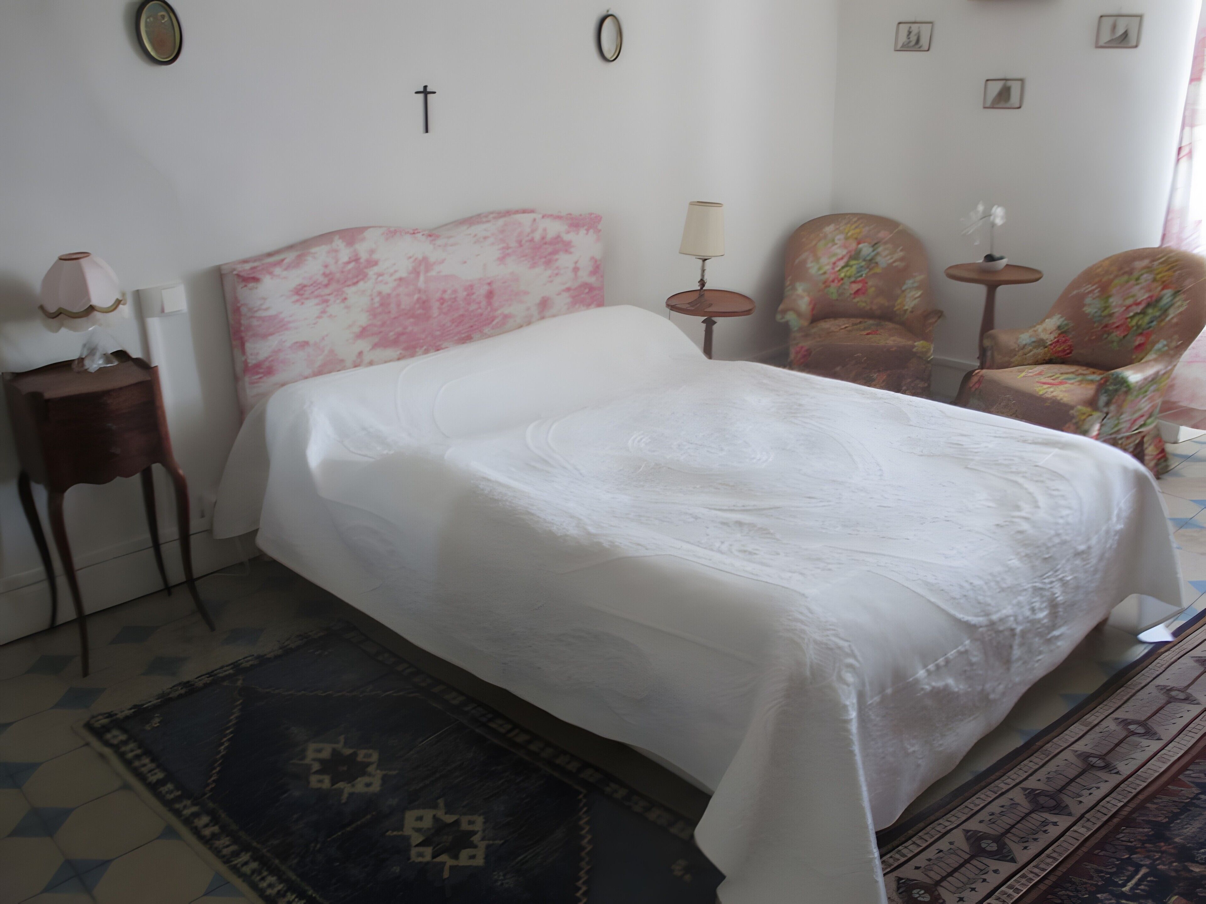 Een bed met een witte spreidekker, een klein nachtkastje met een lamp, een stoel met een bloemenprint en een tafeltje met een plant.