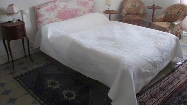 A bed with a white bedspread, a small bedside table with a lamp, a chair with a floral pattern, and a small table with a plant. - Bed and Breakfast 'Chambre Isabeau' with Shared Pool, Shared Garden and Wi-Fi (Carcassonne)