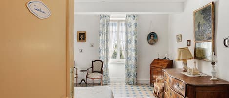 A room with a bed, a wooden dresser, a chair, and a table with a lamp. There is a window with curtains and a door with a nameplate.