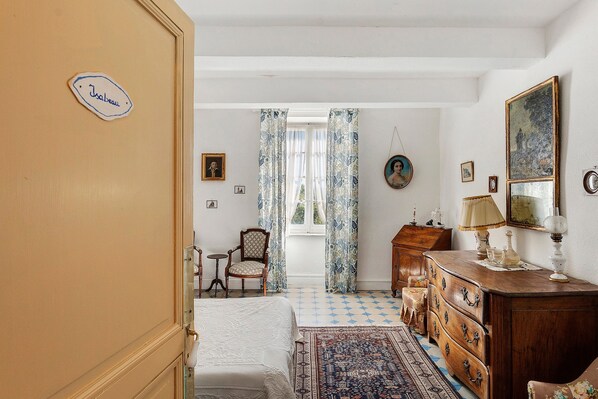 A room with a bed, a wooden dresser, a chair, and a table with a lamp. There is a window with curtains and a door with a nameplate.