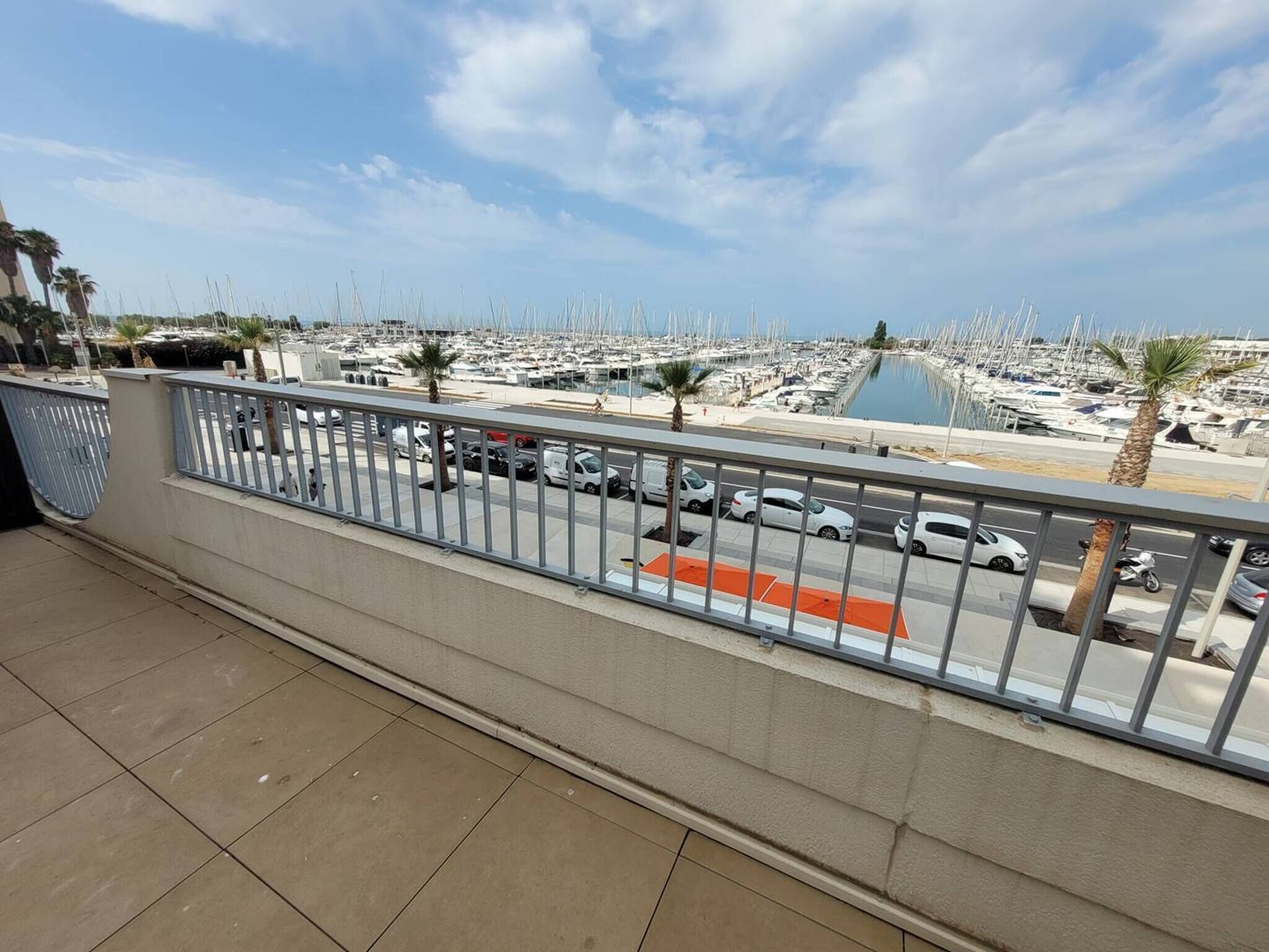 Un balcon donnant sur une marina remplie de bateaux, avec un ciel dégagé et quelques palmiers.