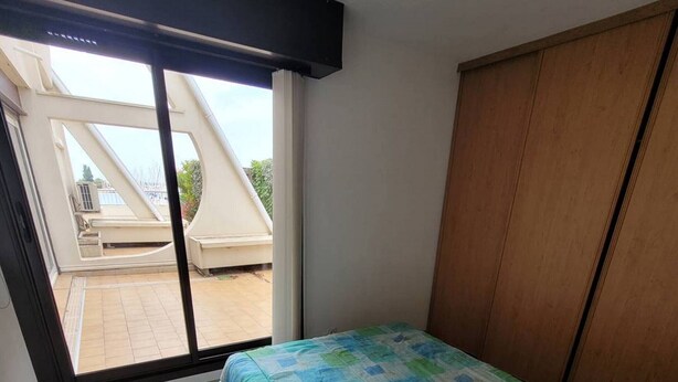 A bedroom with a large window, a bed with a patterned bedspread, and a wooden wardrobe.