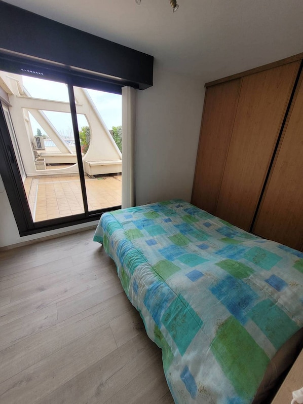 A bedroom with a large window, a bed with a patterned bedspread, and a wooden wardrobe.