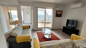 A modern living room with a sofa, a coffee table, and a television. There is a sliding glass door leading to a balcony with a city view. - Quartier du port, t4 pour 8 personnes N12 (La Grande-Motte)