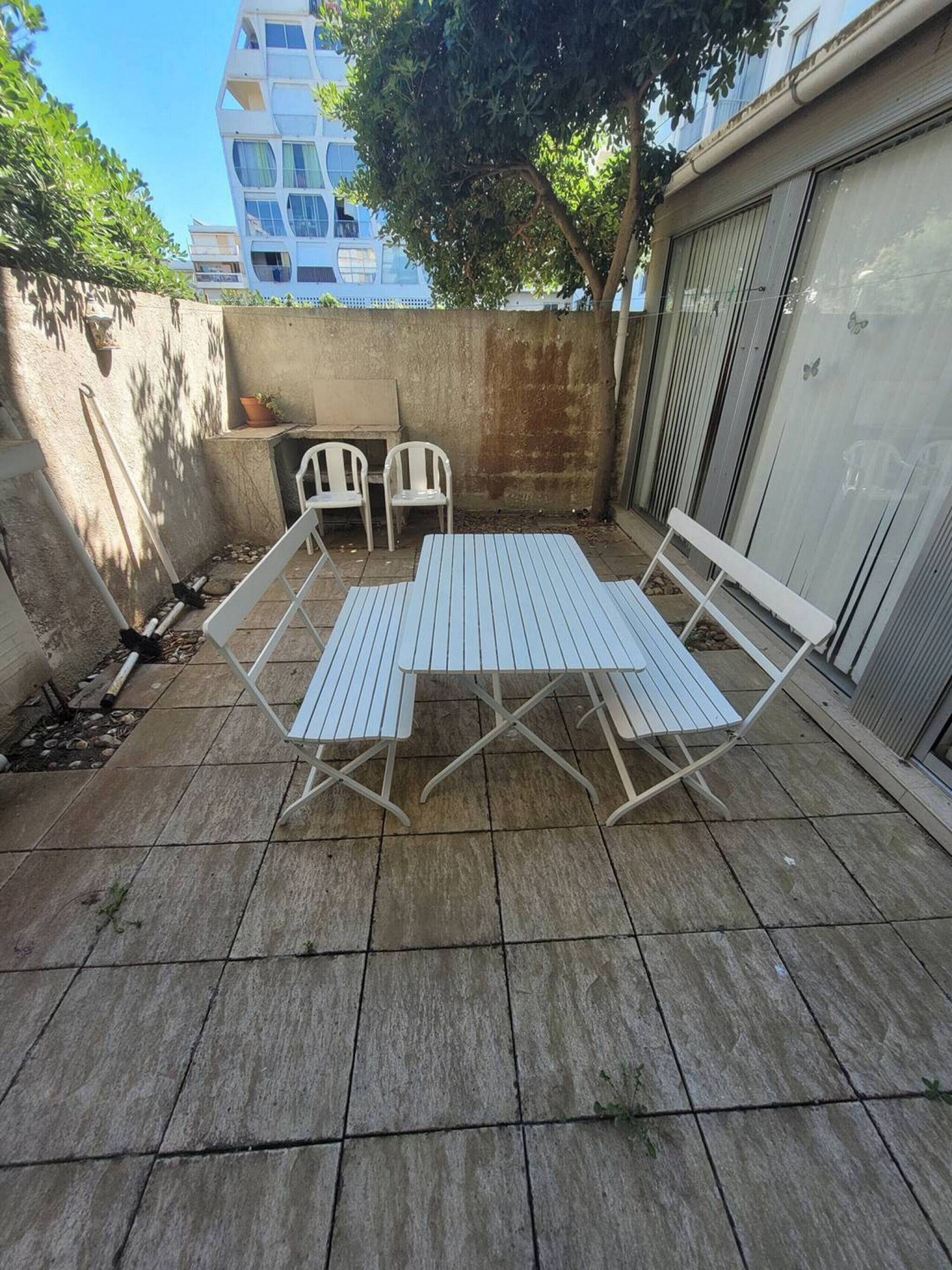 An outdoor patio with a metal table and four chairs, a concrete floor, and a building in the background.