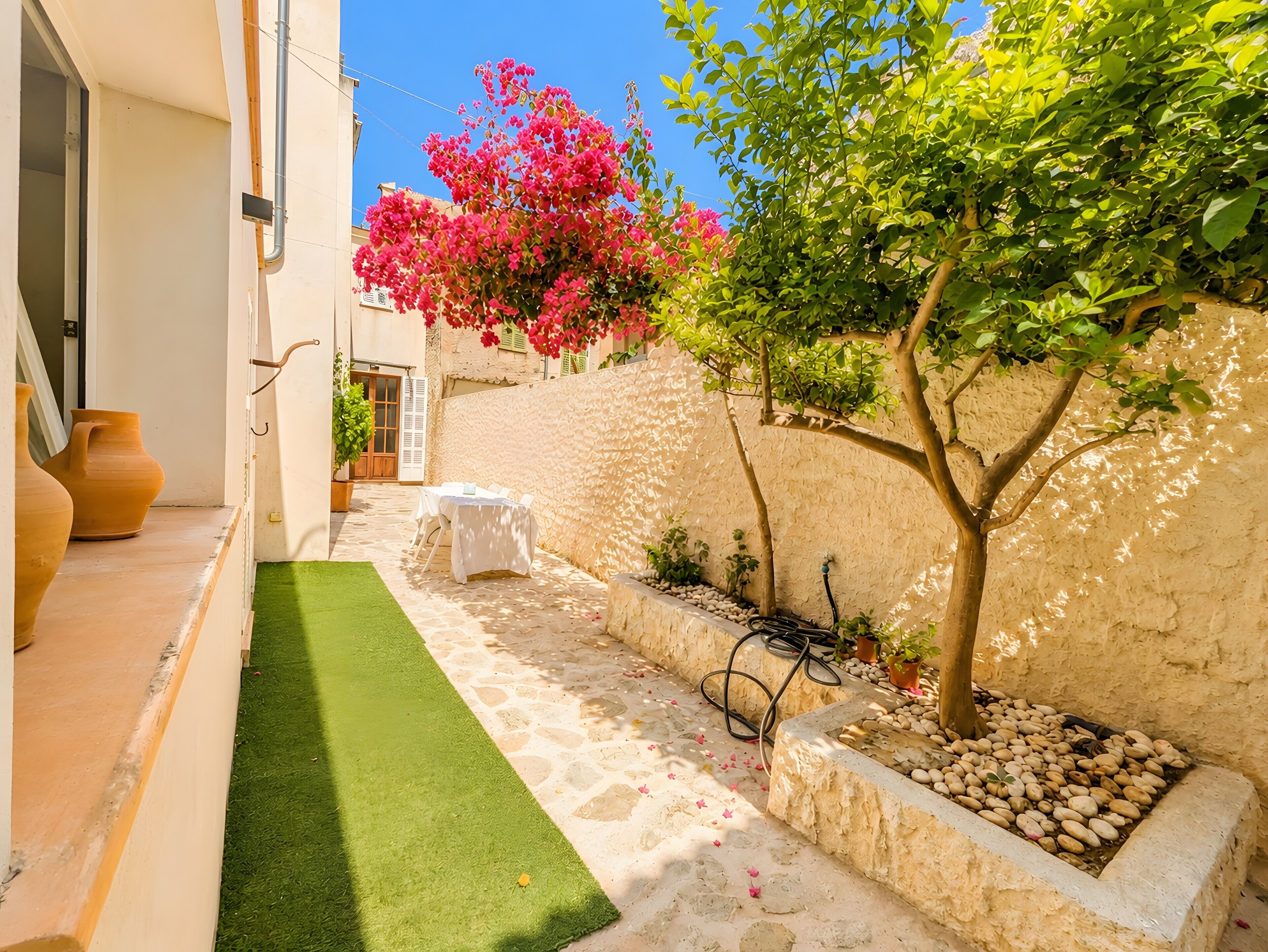 A narrow outdoor space with a white wall, a small tree, a white table, and a green artificial turf.