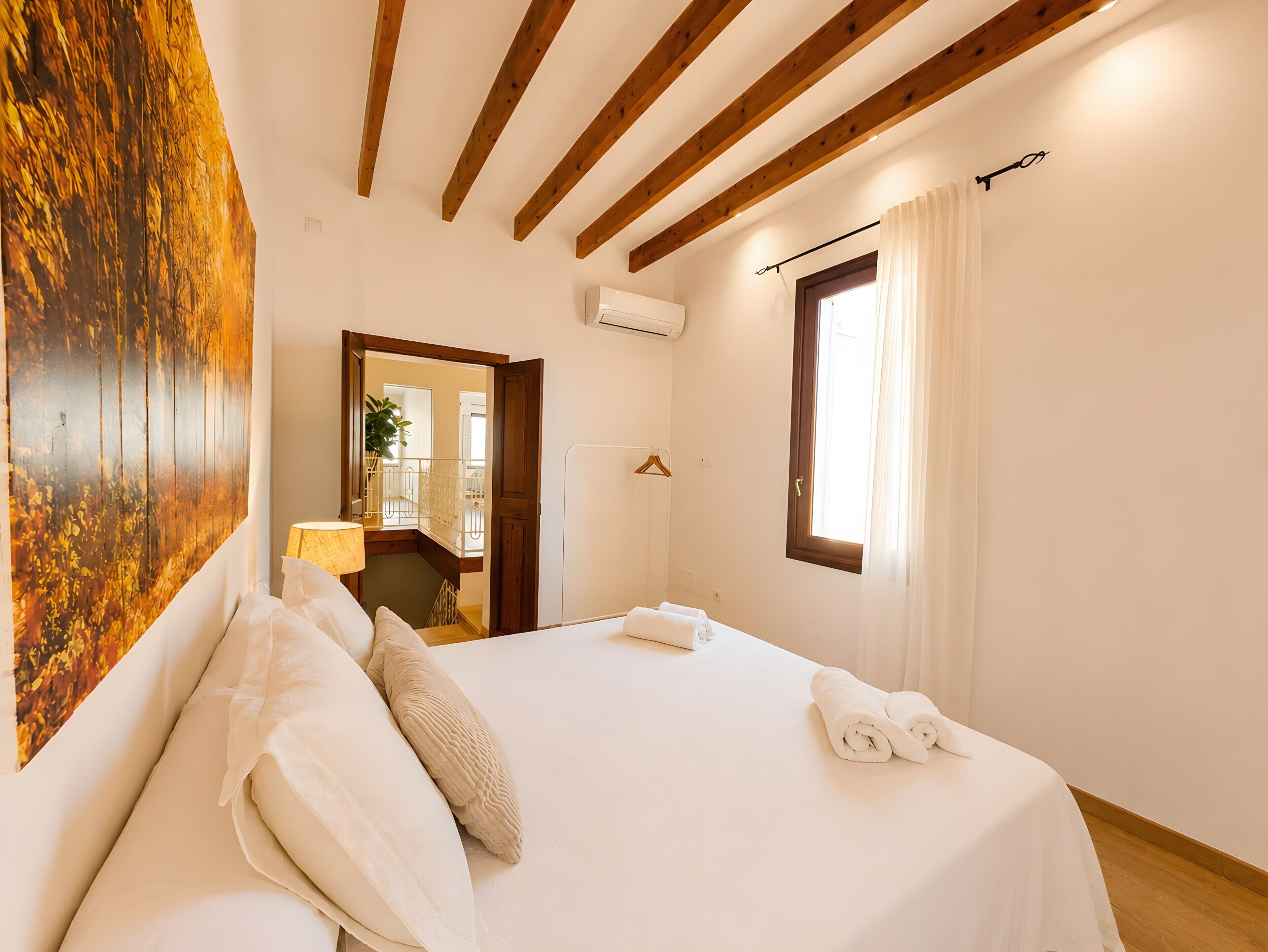 A hotel room with a bed, a painting, a window with curtains, and a wooden ceiling.