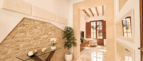 A spacious hallway with a stone wall, a glass table with decorative items, a potted plant, and a view of a room with a window.