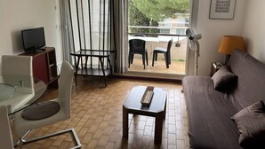 Una camera d'albergo con divano, un piccolo tavolo, una televisione, una sedia, un balcone con tavolo e sedie e una finestra con tende.