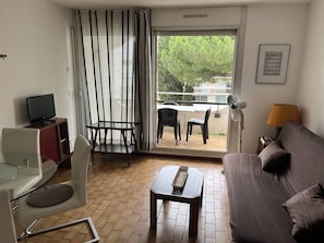 Habitació d'hotel amb sofà, taula petita, televisió, cadira, balcó amb taula i cadires, i finestra amb cortines.
