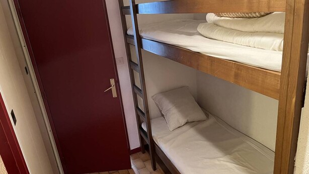 Un letto a castello con struttura in legno e biancheria bianca, una porta rossa e un pavimento piastrellato.