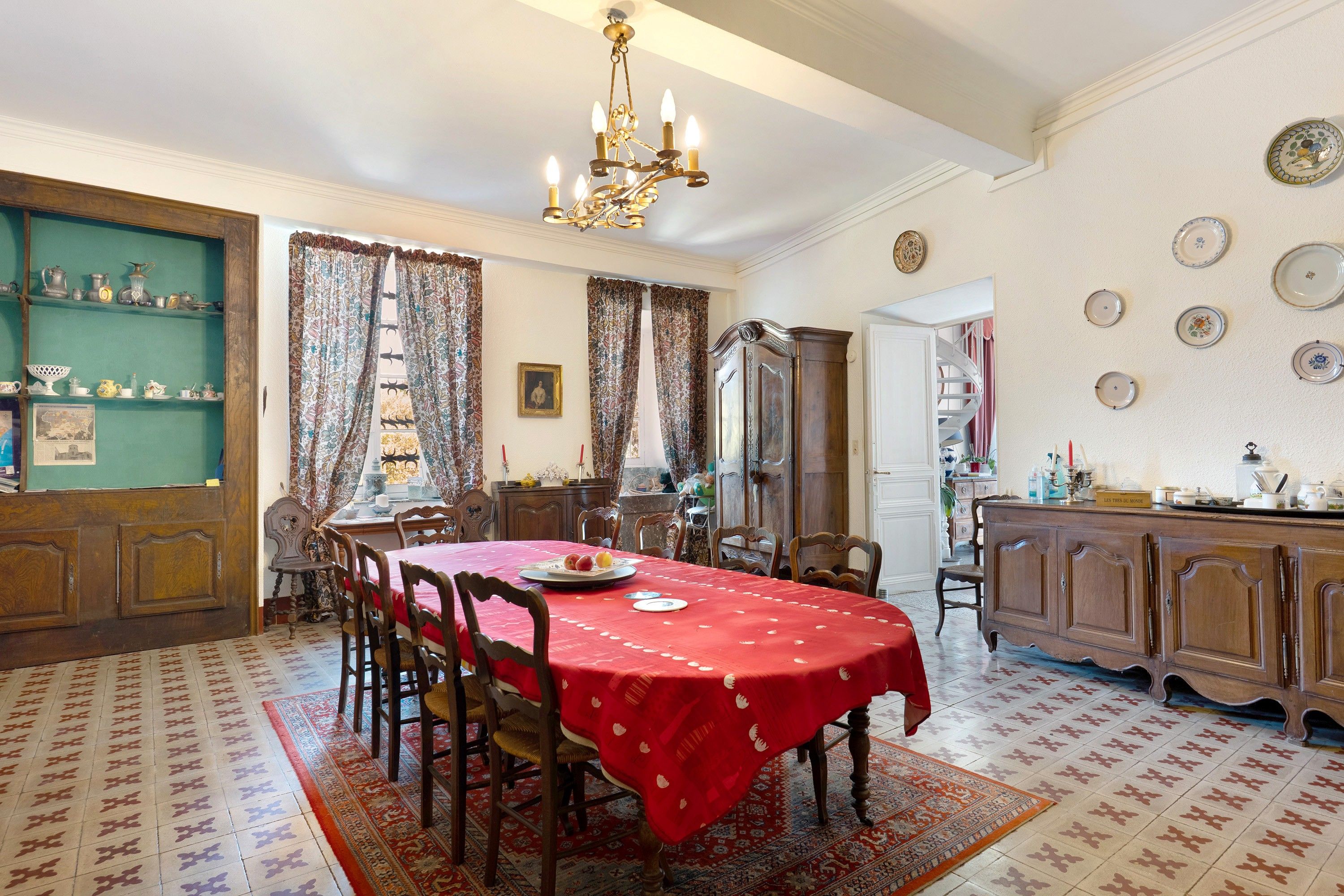 A dining room with a red tablecloth, wooden chairs, a chandelier, and a patterned rug.