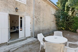 A rustic building with a door leading to a bedroom, a small outdoor seating area with a table and chairs, and a garden with trees and shrubs.
