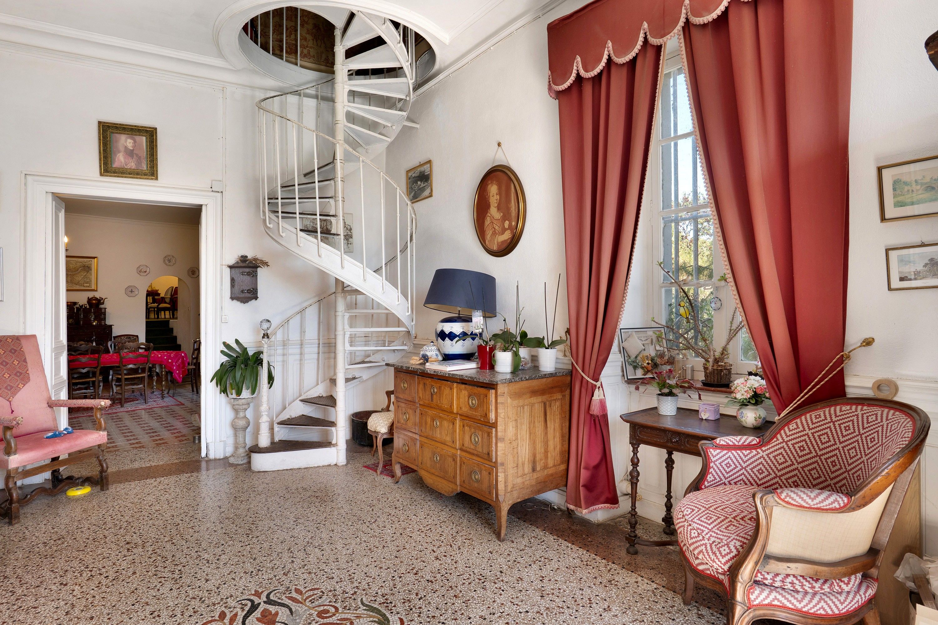 A room with a spiral staircase, a wooden cabinet, a patterned chair, and a dining area with a red tablecloth.