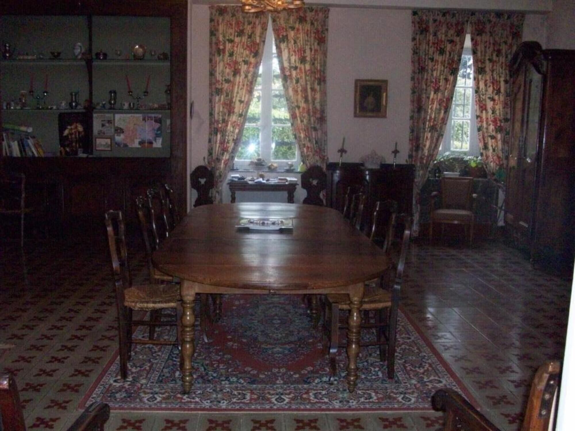 Ein Esszimmer mit einem runden Holztisch, umgeben von Stühlen, einem gemusterten Teppich und einem Bücherregal mit verschiedenen Gegenständen.