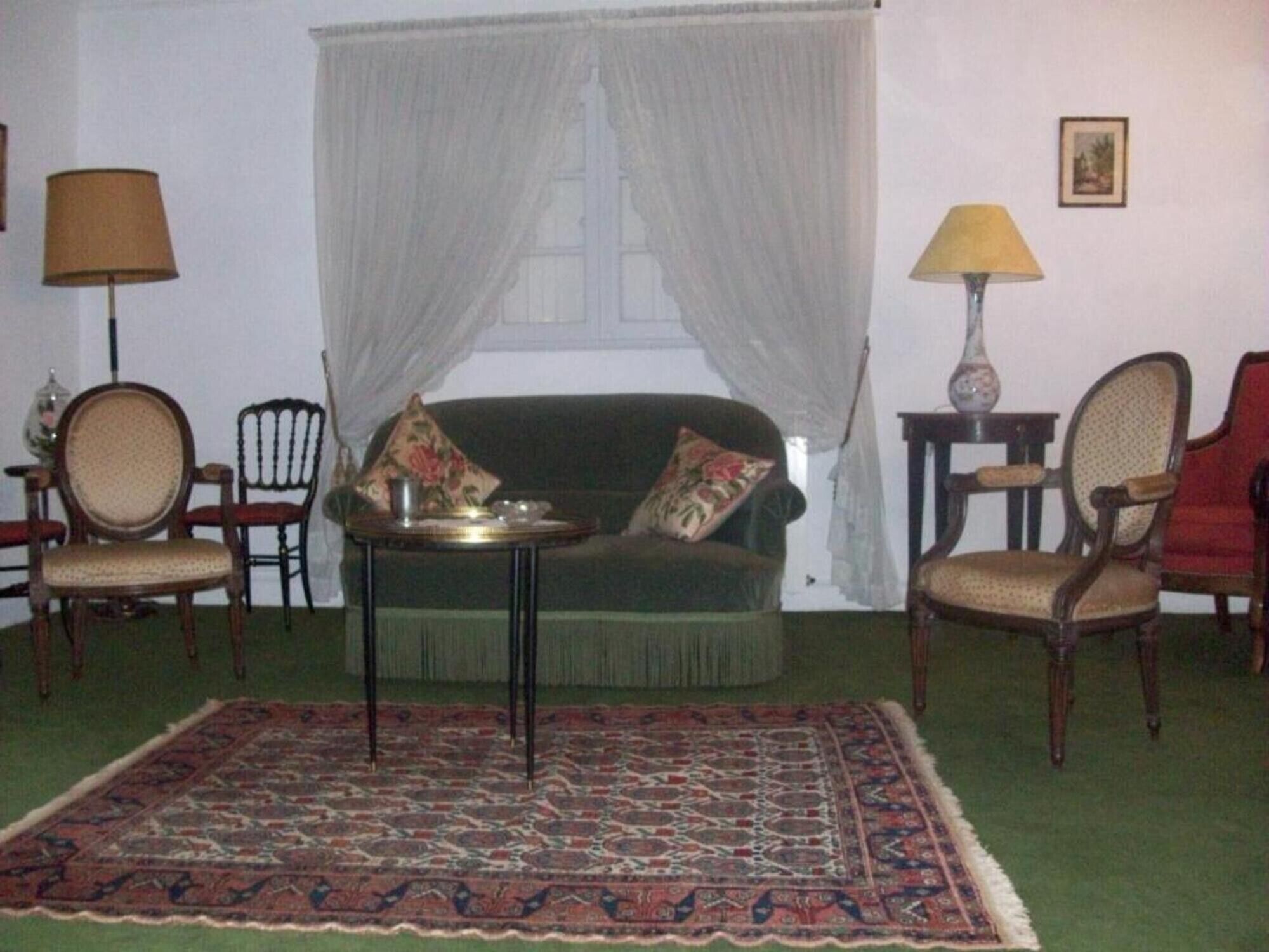 Een kamer met een groene bank, twee fauteuils, een kleine tafel, een kamerkleed met dessin, een raam met dunne gordijnen en twee lampen.
